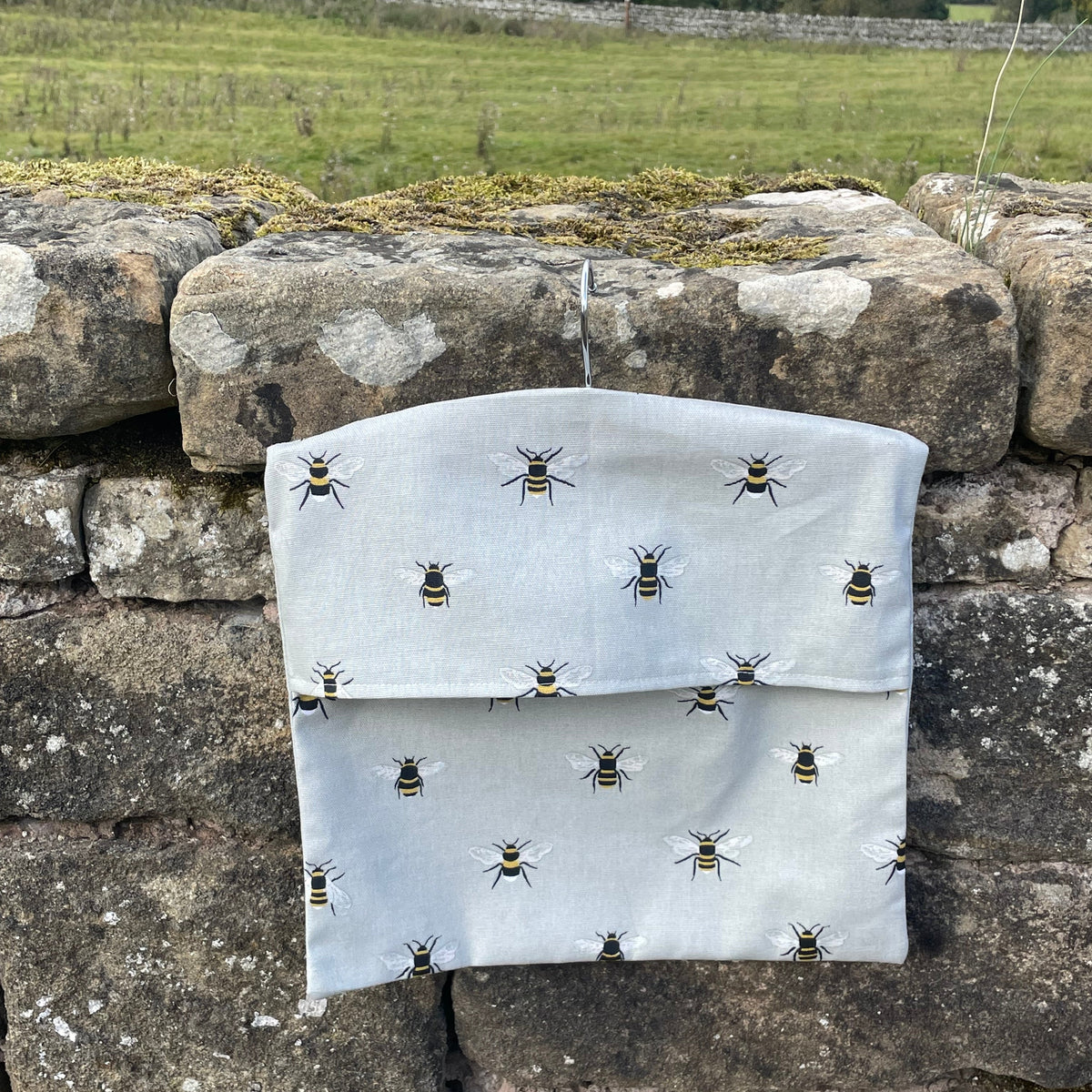 A light gray fabric pouch with a repeating bee print, hanging on a rustic stone wall with a grassy field in the background.