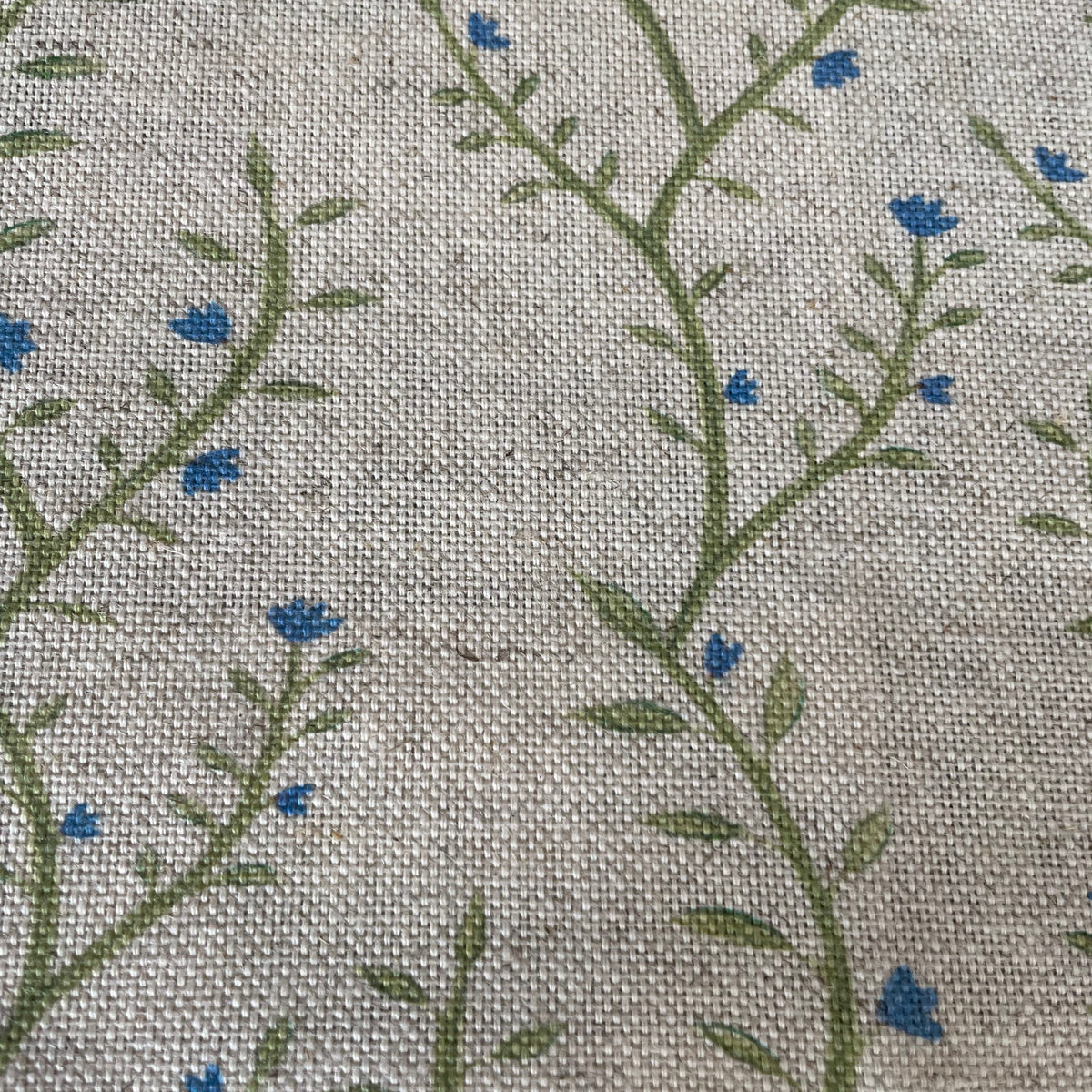 Close-up of a woven fabric with a repeating pattern of green leafy vines and small blue flowers on a light beige background.