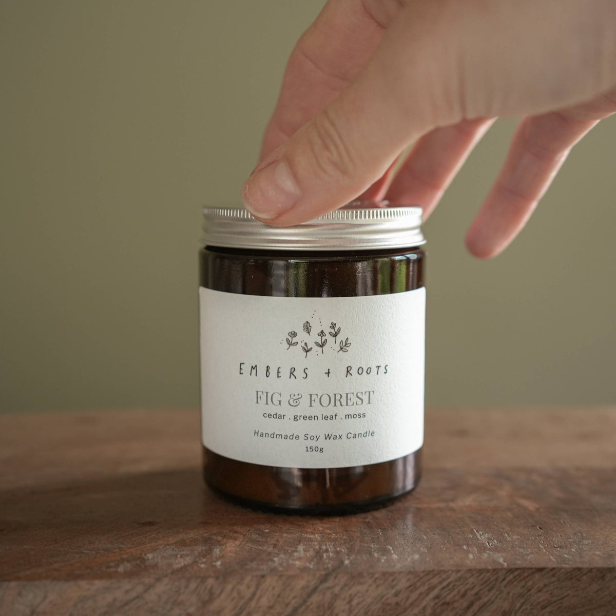 A hand is lifting the lid of an amber glass jar candle on a wooden surface. The candle has a white label that reads 'Ember's + Roots Fig & Forest' and a silver metal lid.